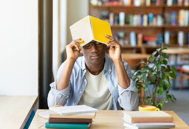 Przepracowany czarny facet śpiący podczas studiów w miejskiej kawiarni, zakładający książkę na głowę, zmęczony robieniem domowych zadań. - Zdjęcie, obraz