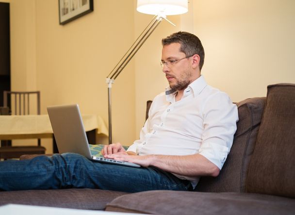 Jeune homme en lunettes avec ordinateur portable sur canapé à l'intérieur de la maison
  - Photo, image
