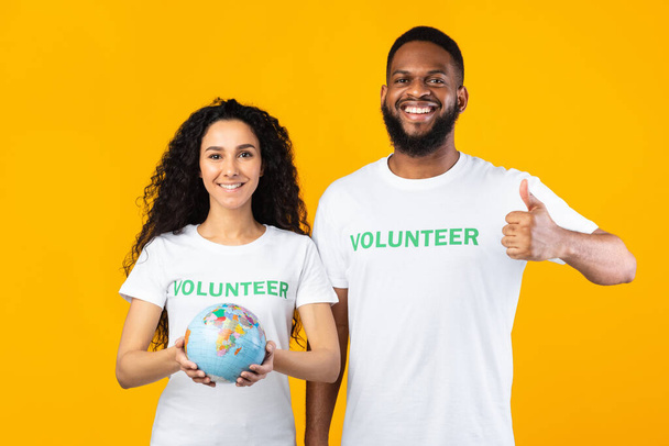 Két különböző önkéntesek Holding Globe Gesturing Thums-Up, Sárga háttér - Fotó, kép
