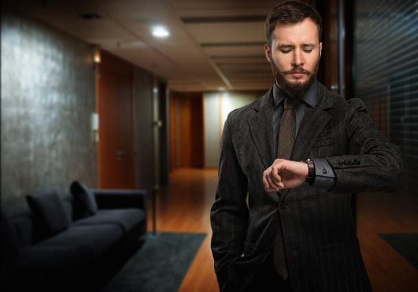Handsome well-dressed man with beard looking at his wrist watch in a hallway - Photo, Image