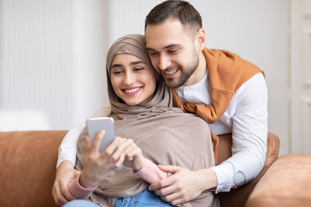 Happy Muslim Couple Using Smartphone Texting Sitting On Couch Indoors - Photo, Image