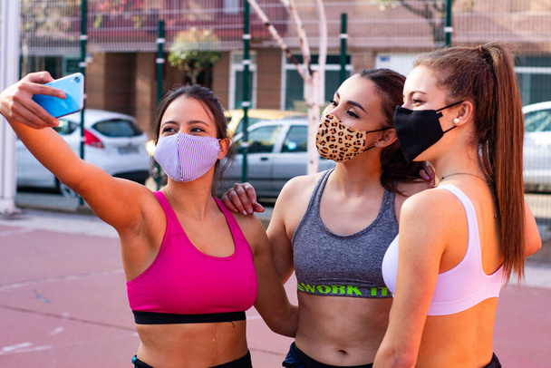 Tres chicas alegres con máscaras se toman una selfie mientras practican baloncesto en una cancha de la ciudad - Foto, Imagen