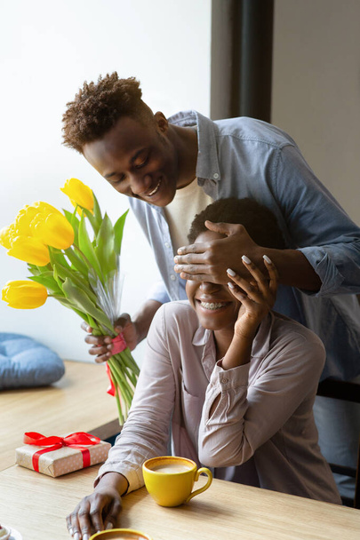 Kochający Afroamerykanin prezentuje swoją dziewczynę z żółtymi tulipanami, zakrywając jej oczy, robiąc niespodziankę w kawiarni - Zdjęcie, obraz