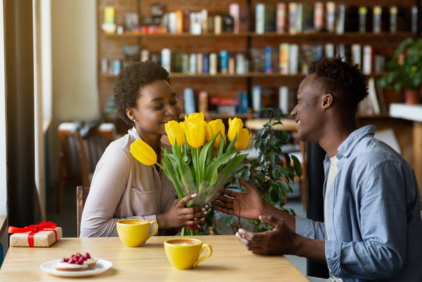 Поганий чорний хлопець дарує своїй дівчині букет тюльпанів і подарунок за Валентинів день у кафе. - Фото, зображення