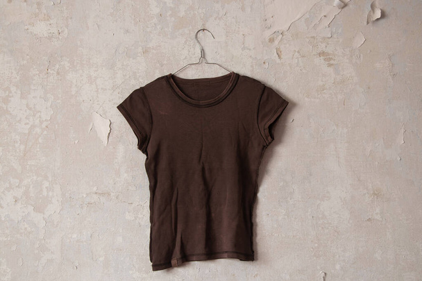 καφέ γυναικείο t-shirt κρέμεται σε μια κρεμάστρα σε ένα παλιό βρώμικο λευκό τοίχο στο σπίτι - Φωτογραφία, εικόνα