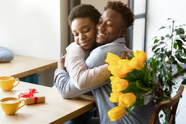 黄色の花と贈り物が一緒にバレンタインデーを祝うカフェで互いに抱き合っている黒いカップルを愛情を込めて - 写真・画像