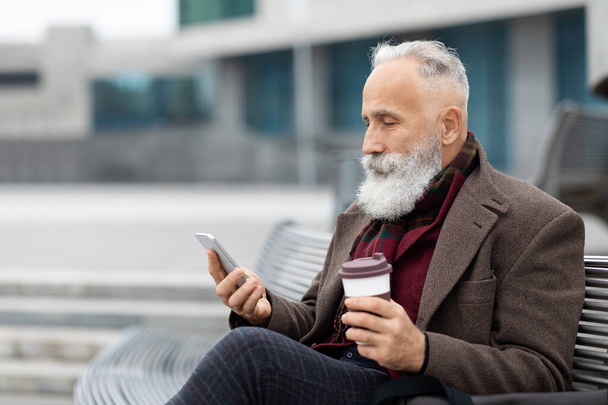 Hombre mayor de pelo gris en traje cálido usando teléfono móvil al aire libre - Foto, imagen
