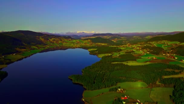 Beau paysage culturel nordique avec lac bleu clair, champs verts et fermes et montagnes enneigées au loin. - Séquence, vidéo