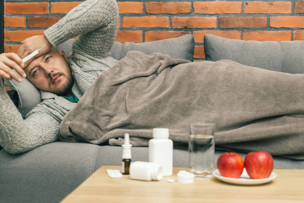 Beteg ember kanapén méri a testhőmérsékletet, ellenőrizze a hőmérőt, a fickó beteg és rosszul érzi magát, fertőzés coronavirus, covid-19 vagy influenza betegség. - Fotó, kép