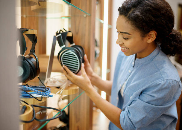 Woman holds vintage headphones in audio components store. Female person in music shop, showcase with earphones on background, buyer in multimedia salon - Photo, Image