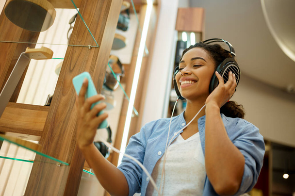 Woman listening to music in headphones store. Female person in audio shop, showcase with earphones on background, buyer in multimedia salon - Photo, Image