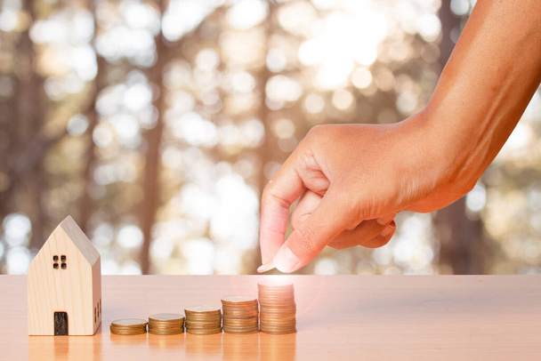 Geld sparen Hand legt Münzen auf einen Stapel mit einem hölzernen Modell zu Hause auf unscharfem Blattgrund. Konzept für den Hauskauf. - Foto, Bild