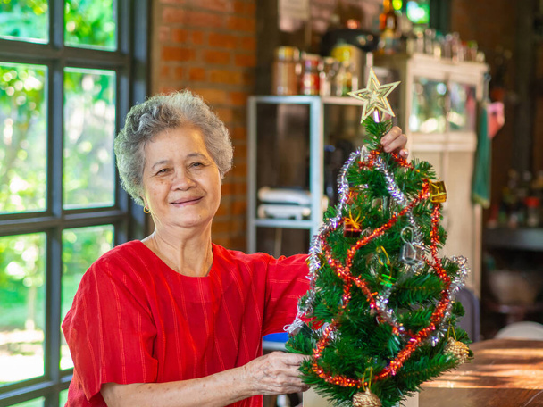 Mujer mayor decorando el árbol de Navidad con cintas blancas y rojas mientras está sentada en una silla en casa. Concepto de personas mayores y festival de Navidad y año nuevo. - Foto, imagen