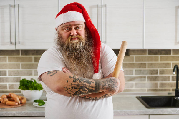 Un homme en surpoids au chapeau de Père Noël avec des bras tatoués croisés et un rouleau à pâtisserie sur sa main regardant la caméra montrant ainsi son émotion - l'indifférence - Photo, image