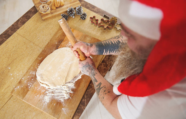 A top view of a man holding a wooden rolling pin like a baseball bat. A flat dough and some Christmas decorations on a table in the kitchen - Photo, Image