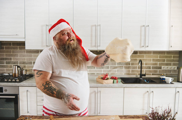 Щасливий бородатий чоловік у капелюсі Санта Клауса і татуювані руки, які готують тісто і викидають його, як кваліфікованого шеф-кухаря на домашній кухні. Готування різдвяного печива - Фото, зображення