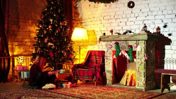 Χριστουγεννιάτικη διάθεση μιας νεαρής γυναίκας που κάθεται σε ένα άνετο σαλόνι με τζάκι και βάζει δώρα - Πλάνα, βίντεο