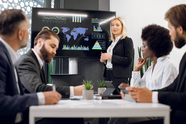 Middle aged pleasant confident blond business lady, showing companys work infographics on digital wall screen during meeting with team of diverse multiracial male and female businesspeople - Foto, imagen