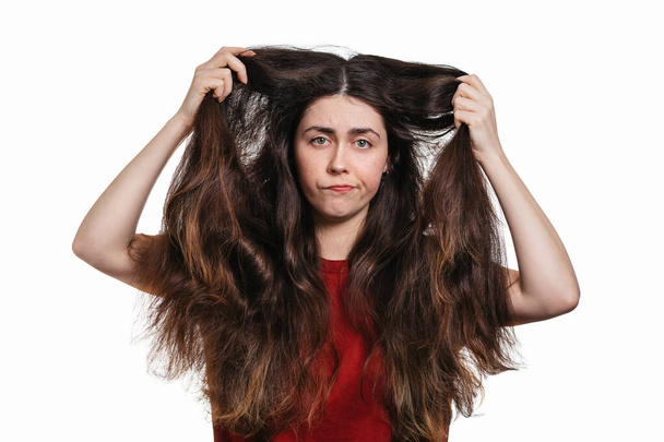 Portrét mladé nespokojené ženy ukazuje její husté a nepoddajné vlasy. Bílé pozadí. Koncept barvení a péče o vlasy. - Fotografie, Obrázek