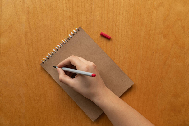 白紙のクラフト紙のノートにメモを取る。木製のテーブルの上の女性の手と赤い鉛筆の背景。新年の抱負や目標です。2021年コロナウイルス。ロックダウン、割り当て、ホームオフィス. - 写真・画像