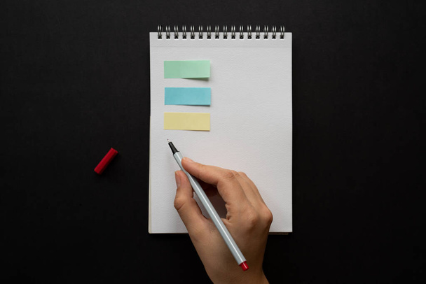 白いノートにカラフルなポストイットの女性の手と木製のテーブルの背景に赤い鉛筆でメモを取る。新年の抱負や目標です。2021年コロナウイルス。自宅のオフィスでリストを作成しスペースをコピーし. - 写真・画像