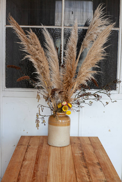 dry flowers and fluffy reeds in a beige ceramic vase on wooden table against a white wall with dark window - Фото, изображение