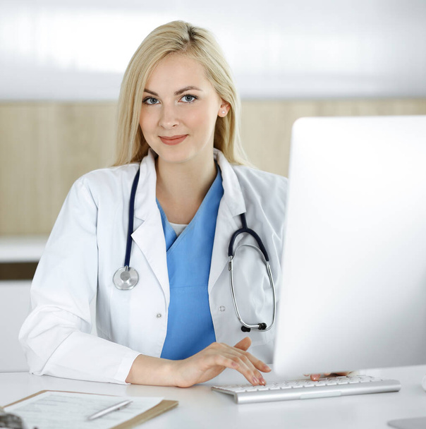 Woman-doctor at work while sitting at the desk in hospital or clinic. Blonde cheerful physician using desktop computer - Foto, afbeelding