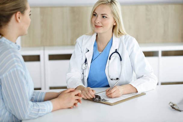Woman-doctor and patient sitting and talking in hospital or clinic. Blonde therapist is cheerfully smiling. Medicine concept - Фото, зображення