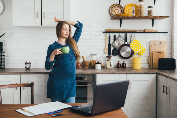 Young pretty pregnant freelancer girl works at home in the kitchen in self-isolation mode in quarantine and does a warm-up from fatigue. Toning. - 写真・画像
