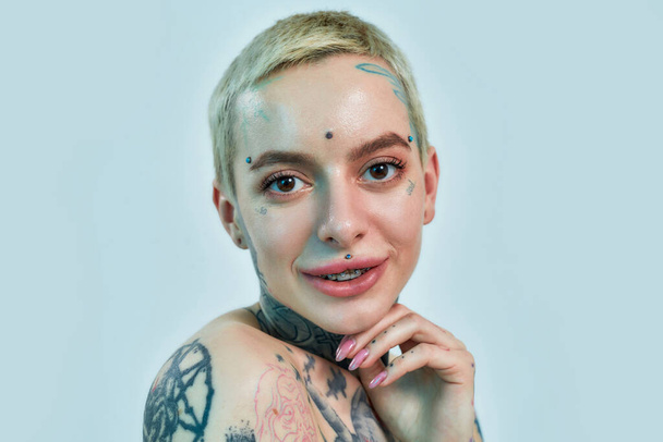  Close up of a white tattooed woman with piercing and brackets looking into a camera on a blue background - Zdjęcie, obraz