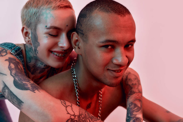 Young white pierced tattooed female with eyes closed hugging dark-skinned topless tattooed male from back , both smiling on a bright pink background - Foto, imagen