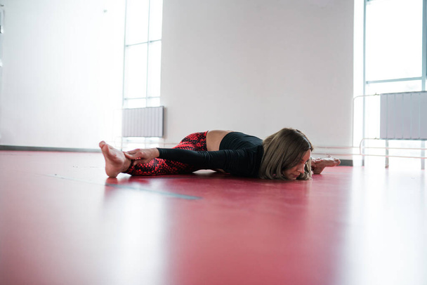 A girl stretching trainer lies in a transverse twine on a red gymnastic mat in a light fitness studio. - Φωτογραφία, εικόνα