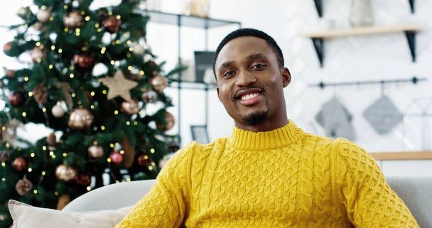 Cheerful African American male in good mood looking at camera and smiling indoors near christmas glowing decorated tree with lights and feeling happy on Xmas eve Merry christmas Portrait concept - Foto, immagini
