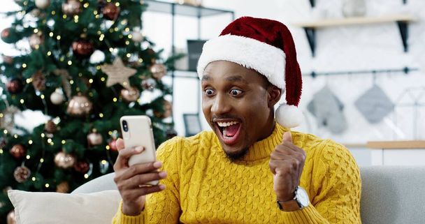 Афроамериканец радостно удивленный человек в канун Рождества получает отличные новости по мобильному телефону о победе лотереи на аукционе, чувствуя себя счастливым. Невероятная удача, новогоднее чудо, искренние эмоции, рождественская распродажа - Фото, изображение