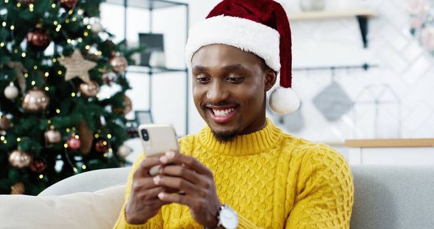 Close up of handsome African American man sitting on sofa in modern home near Christmas glowing tree and texting on smartphone sending holiday greetings to friends. New Year concept - Foto, imagen