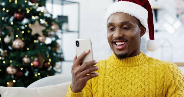 Close up of handsome African American man sitting on sofa in modern home near christmas glowing tree and texting on smartphone sending holiday greetings to friends. Best wishes. New Year concept - Фото, изображение