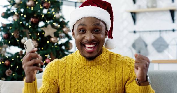 Primer plano de afroamericano alegre sorprendido hombre emocionado recibir grandes noticias en el teléfono inteligente acerca de ganar la lotería en la víspera de Navidad Guy sentirse feliz de tener suerte increíble emociones felices ventas de Año Nuevo - Foto, imagen