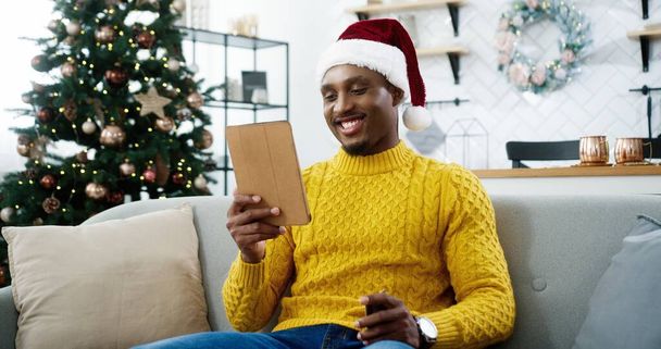 Close up of happy African American male shopper in santa hat sitting in decorated home near christmas glowing tree and buying online on tablet paying with credit card on xmas sales. E-commerce concept - Foto, imagen