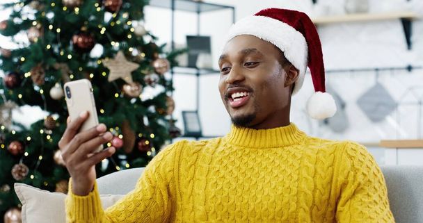 Close up of handsome guy in xmas hat videochatting on cellphone while sitting near decorated new year tree at home. Cheerful African American man smiling and speaking on video chat. New Year concept - Photo, Image