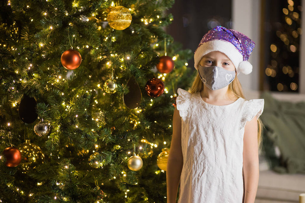 Niña en máscara protectora y sombrero de santas en la habitación de Navidad decorada. - Foto, Imagen