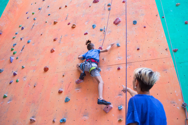 Ein Trainer unterrichtet ein Kind, eine Frau begleitet ihren Sohn, ein Junge und seine Mutter trainieren zusammen, Kinder klettern, ein Junge klettert die Wand hinauf, Kletterhalle - Foto, Bild