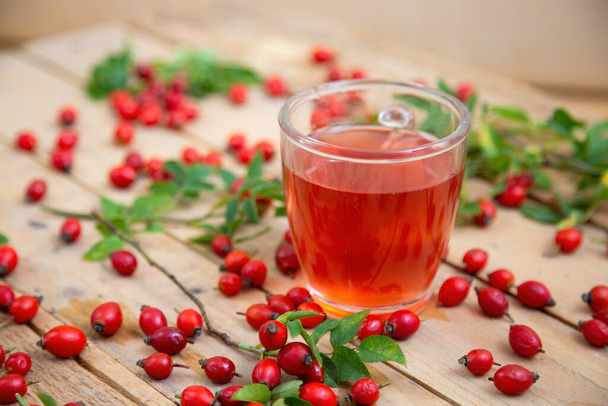 Glass of rosehip tea on brown palette with leaves and berries around - 写真・画像