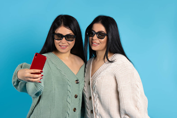 Kaksoset 3D-lasit, samoissa vaatteissa, ottavat selfien punaisella älypuhelimella. Sininen tausta. Laadukas kuva - Valokuva, kuva