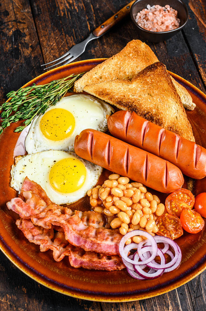 Tradiční anglická snídaně se smaženými vejci, klobásami, slaninou, fazolemi a tousty. Tmavé dřevěné pozadí. pohled shora. - Fotografie, Obrázek