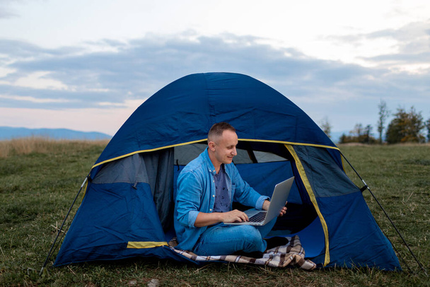 Man working on laptop in tent in nature. Young freelancer sitting in camp. outdoor activity in summer. internet 5G - Фото, изображение