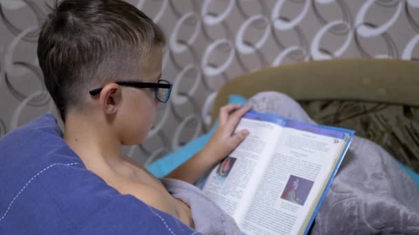 Serious Boy avec des lunettes lit un livre intéressant à s'asseoir sur le lit dans la couverture - Séquence, vidéo