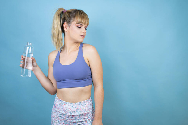 Молодая красивая женщина в спортивной одежде держа бутылку воды на изолированном синем фоне серьезно и глядя на сторону - Фото, изображение