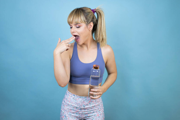 Spor kıyafetleri giyen genç bir kadın bir şişe su tutarken izole edilmiş mavi arka planda eli ağzında. - Fotoğraf, Görsel