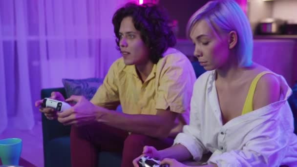 Boldog Y-generációs pár, akik joystickokat tartanak, beszélgetnek, videojátékoznak, nevetnek a kanapén. Mosolygó férfi és nő együtt töltik az időt otthon. - Felvétel, videó