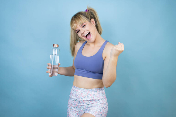 Jeune belle femme portant des vêtements de sport tenant une bouteille d'eau sur fond bleu isolé très heureux et excité faisant geste gagnant avec les bras levés, souriant et criant pour le succès. - Photo, image